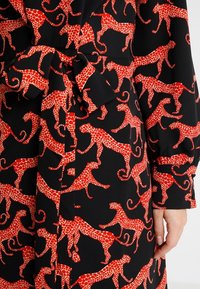Schwarzer Stoff mit einem sich wiederholenden Muster von roten Leoparden mit gepunkteten Details. Inkludiert einen Bindegürtel in der Taille und lange Ärmel.