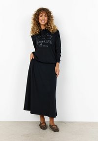 Čierna mikina s kapucňou a textom, dlhé rukávy a klokanie vrecko, v kombinácii s dlhou, splývavou čiernou sukňou a hnedými slip-on topánkami s detailmi.