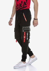 Schwarze Cargo-Hosen mit roten Akzenten, mehreren Reißverschluss-Taschen und Nieten. Eng anliegende Bündchen an den Knöcheln, kombiniert mit weißen Sneakers.