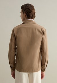 Camicia marrone in tessuto intrecciato con maniche lunghe, colletto e polsini con bottoni, caratterizzata da un taglio dritto e senza motivi visibili o dettagli metallici.