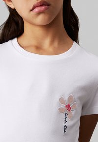 Camiseta de algodón blanca con cuello redondo. Presenta un diseño de bordado floral en suave rosa y rojo con texto en azul oscuro que dice "Calvin Klein."