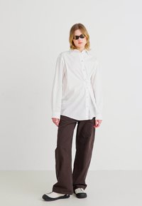 White button-up shirt with knotted front detail, paired with loose-fitting brown pants and black sunglasses. Textured fabric throughout.