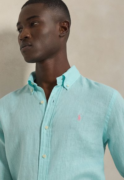 Camisa azul claro de lino con botones y un detalle de logo rosa, diseño de cuello clásico, mostrada en la parte superior del cuerpo de un modelo.