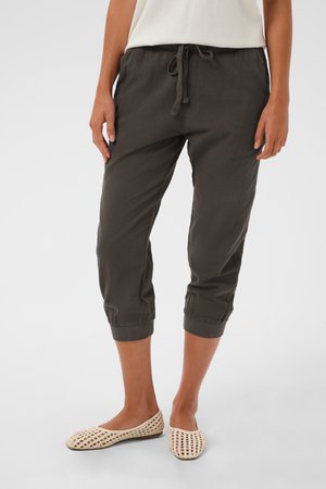 Person iført mørk grå korte bukser med snor i taljen og beige vævede slip-on sko, der står på en ensfarvet baggrund.