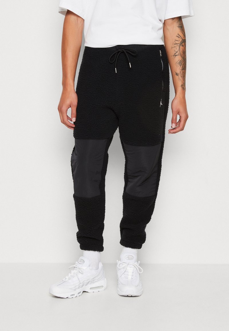 Schwarze Fleece-Jogginghosen mit glatten, schwarzen Nylon-Knieteilen, elastischem Bund mit Kordelzug, Seitentaschen und einem Markenakzent am Oberschenkel.