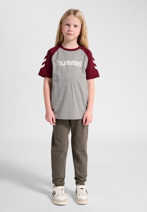 Jong meisje staand, draagt een grijs- en bordeauxrood 'hummel' t-shirt, olijfgroene broek en witte sneakers met zwarte strepen.