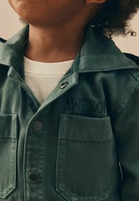Veste en coton verte avec un col, une fermeture à boutons et deux poches poitrine. Présente le mot "AWESOME" en noir sur le côté gauche.