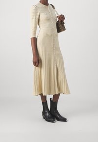 Strickad cremefärgad klänning med strukturerade mönster, knappad front och två fickor; i kombination med svarta ankelstövlar med fyrkantig tå och låg klack.