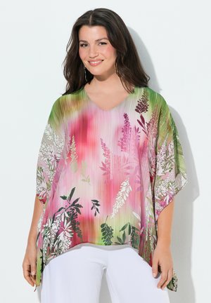 Uśmiechnięta kobieta w luźnej bluzce w kolorowy kwiatowy wzór w odcieniach różu, zieleni i bieli, w połączeniu z białymi spodniami.