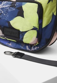 Mała torebka w kwiatowy wzór w kolorach niebieskim, zielonym i czarnym, z czarnym odpinanym paskiem z srebrnym zapięciem oraz naszywką z logo Desigual.