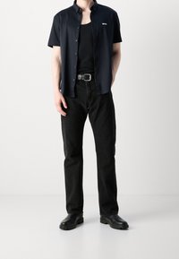 Marineblauwe short-sleeve button-up shirt, zwart tanktop, zwarte jeans en zwarte laarzen. Het shirt heeft een logo op de borst. Klassieke pasvorm en een schoon ontwerp.