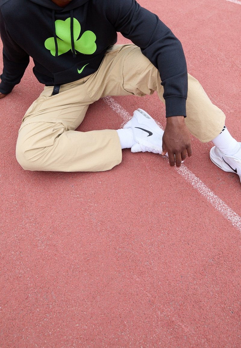 Personne assise sur une piste rouge portant un pantalon beige, un sweat à capuche noir avec un motif de trèfle vert, des baskets blanches Nike et des chaussettes blanches.