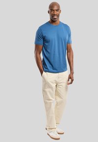 T-shirt en coton bleu à manches courtes, associée à un pantalon cargo crème et des baskets blanches avec des accents beiges. Se tenant devant un fond gris.