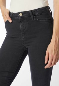 Lähikuva henkilöstä, jolla on mustat skinny-farkut edessä napitus ja käsi taskussa, yksiväristä taustaa vasten.