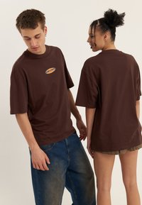 Des t-shirts en coton marron avec un logo rond sur le devant, présentant des manches courtes et une coupe décontractée, sont portés par deux modèles vus sous différents angles.