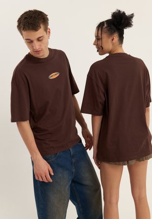 Camisetas de algodón marrón con un logo redondo en la parte frontal, con mangas cortas y un corte relajado, son llevadas por dos modelos desde diferentes ángulos.