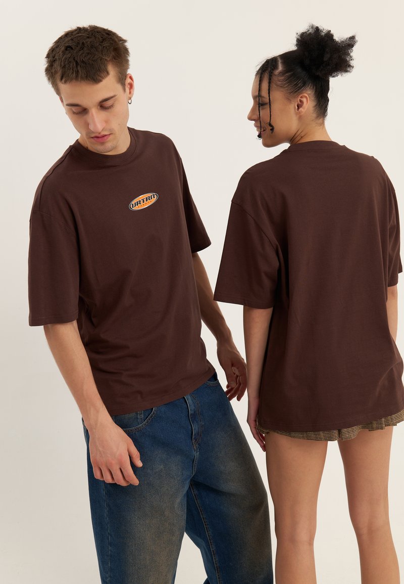 Des t-shirts en coton marron avec un logo rond sur le devant, présentant des manches courtes et une coupe décontractée, sont portés par deux modèles vus sous différents angles.