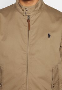 Tanfarbige Jacke mit hohem Kragen, Frontreißverschluss, zwei Seitentaschen und braunen Lederakzenten. Verfügt über ein kleines navyblaues Logo auf der Brust.
