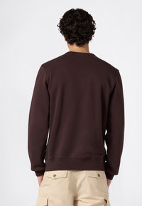 Uomo con capelli ricci scuri che indossa un felpa marrone scuro e pantaloni cargo beige, in piedi voltato di spalle contro uno sfondo bianco semplice.