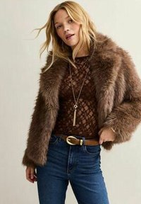 Veste en fausse fourrure marron au fit décontracté, avec un motif texturé, portée par-dessus un haut en dentelle marron. Associée à un jean bleu taille haute et une ceinture.
