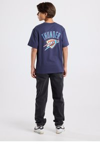 Navy blue short-sleeve T-shirt with "THUNDER" text and logo on back, paired with dark pants and sneakers. Simple design, casual fit.