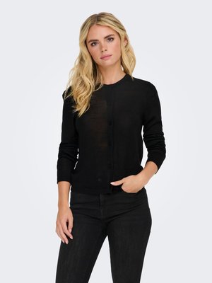 Frau mit langen blonden Haaren, die eine eng anliegende schwarze, geknöpfte Strickjacke und schwarze Hose trägt, steht mit einer Hand in der Tasche vor einem schlichten Hintergrund.