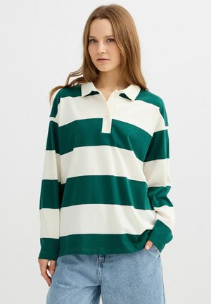 Joven con cabello largo castaño claro, vistiendo una camiseta tipo polo de manga larga con rayas anchas verdes y blancas, y vaqueros azul claro, con la mano en el bolsillo.