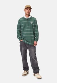 Grün gestreiftes langärmeliges Polo-Shirt mit einem weißen Kragen und Logo auf der Vorderseite, kombiniert mit grauen Jeans und beigefarbenen Sneakers. Akzente sind sichtbare Nähte.