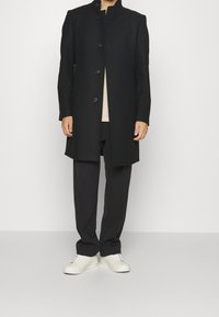 Zwarte lange jas met staande kraag, knoopsluiting aan de voorkant en een zachte textuur. Draagt over een licht shirt en gecombineerd met zwarte broeken en witte sneakers.