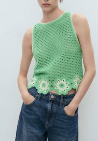 Top smanicato verde realizzato a maglia con motivo a trafori e orlo floreale a onde. Abbinato a jeans di denim blu scuro, mani in tasca.