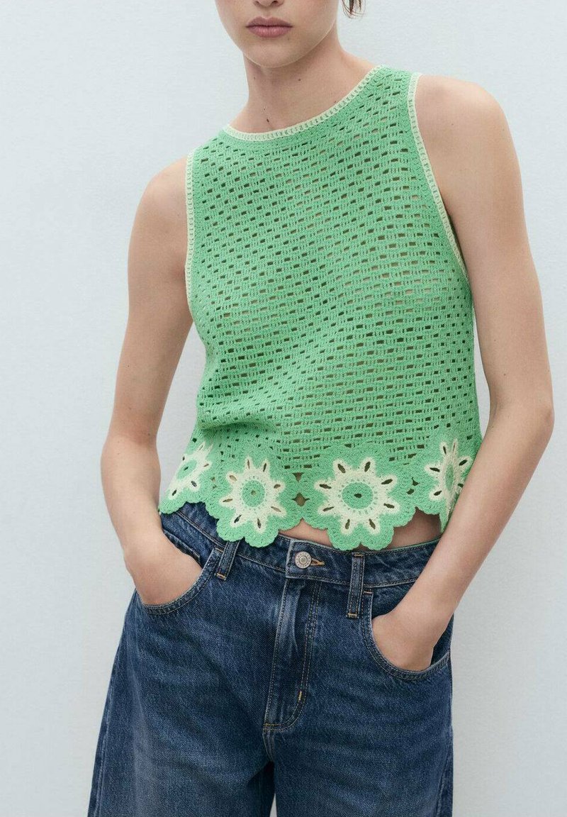 Top smanicato verde realizzato a maglia con motivo a trafori e orlo floreale a onde. Abbinato a jeans di denim blu scuro, mani in tasca.