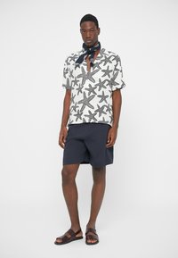 Korte mouwen, overhemd met kraag en zwart zeesterrenpatroon op een witte achtergrond, gecombineerd met marineblauwe korte broek en bruine sandalen. Sjaal om de nek.