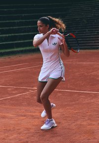 Giocatrice di tennis in abito bianco si prepara a colpire un rovescio su un campo in terra battuta con gradinate verdi sullo sfondo.