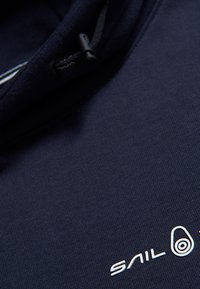 Sudadera azul marino con un tejido texturizado, cordón ajustable y una marca blanca con un logotipo circular en la parte inferior derecha.