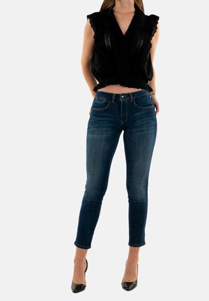 Haut sans manches noir avec des accents en dentelle et un ourlet volant, associé à un jean skinny bleu foncé. Le mannequin porte des talons hauts noirs.