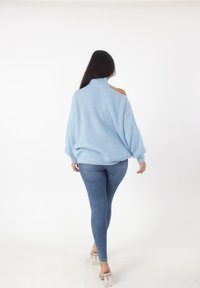 Suéter de punto de gran tamaño en azul claro con un cuello alto y diseño de hombros descubiertos, combinado con jeans ajustados azules y sandalias de tacón transparente.
