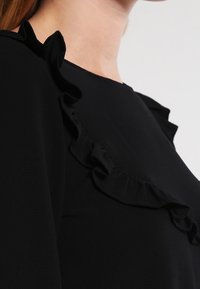 Černý top s řaseným detailem podél výstřihu. Tkanina působí hladce, s jemnou texturou a přiléhavým střihem.