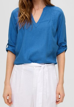 Frau trägt eine blaue V-Ausschnitt-Bluse mit hochgekrempelten Ärmeln und hoch taillierte weiße Hose, die mit einem Stoffgürtel gebunden ist.