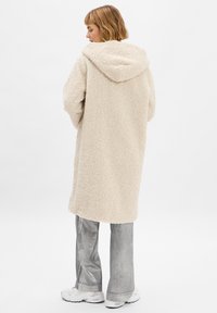 Roomkleurige fleece-jas met een gestructureerd, zacht oppervlak. Gepaard met zilveren, uitlopende broek en witte sneakers.