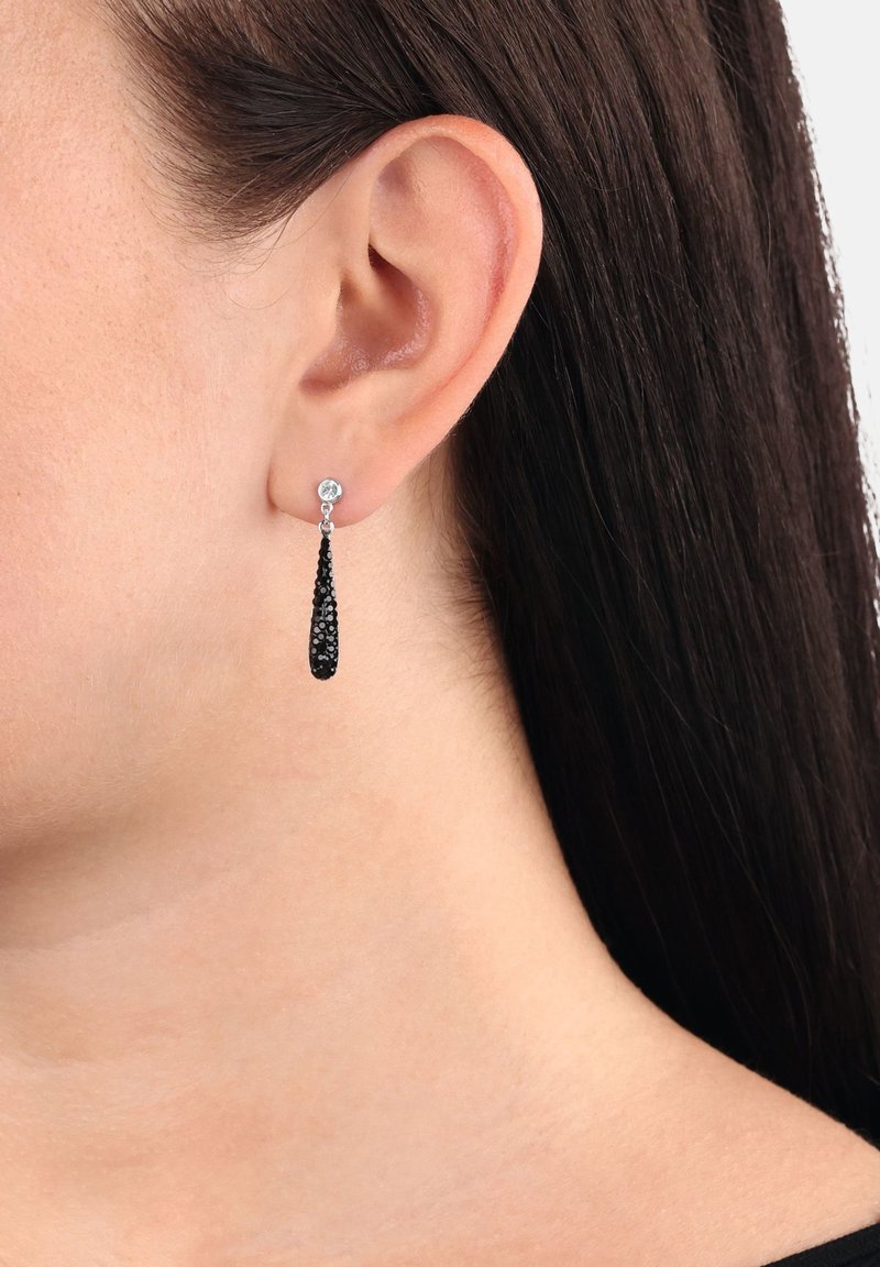 Primer plano de una oreja con cabello lacio y oscuro, que lleva un arete de plata con un colgante negro en forma de lágrima.