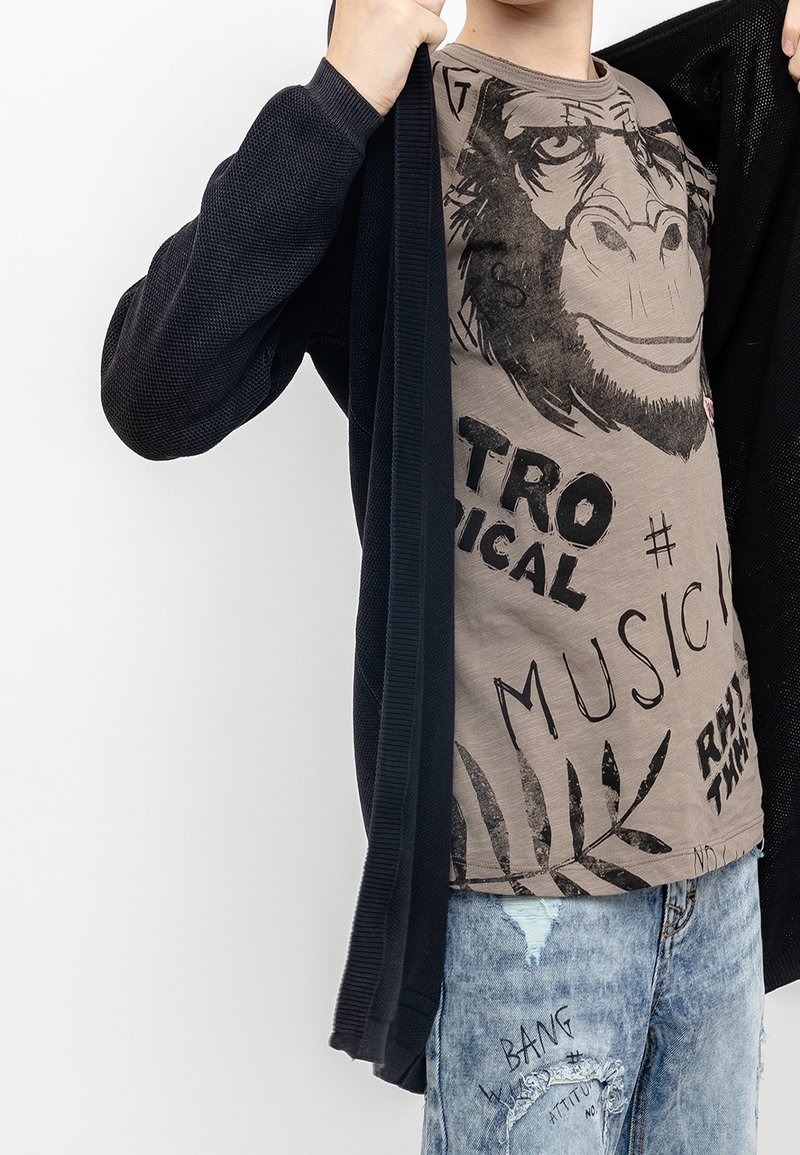 Enfant montrant un t-shirt graphique avec un visage de gorille et du texte, portant un cardigan noir et un jean bleu déchiré avec des motifs imprimés.