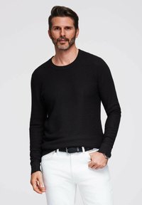 Schwarzer strukturierter Pullover mit langen Ärmeln und Rundhalsausschnitt, kombiniert mit weißen Hosen und einem schwarzen Gürtel. Das Modell posiert mit einer Hand auf der Hüfte.