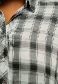 Camicia a quadri bianca e nera con colletto, dotata di chiusura a bottoni e taschino sul petto. Il tessuto appare morbido con una superficie liscia.