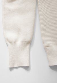 Mansette di un maglione color crema con texture a coste. Una mansetta presenta righe orizzontali, mentre l'altra ha una finitura liscia.