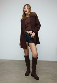 Donna che indossa un cappotto marrone con colletto di pelliccia, top marrone corto, pantaloni shorts in denim scuro e stivali marroni alti fino al ginocchio, in piedi su un pavimento grigio.