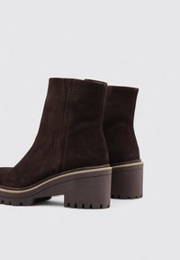 Botas de tobillo de ante marrón con un tacón grueso, que cuentan con una suela texturizada y costuras en contraste a lo largo de las costuras. Acabado suave.