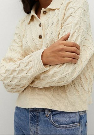 Kvinde iført cremefarvet kabelstrikket sweater med knapper ved kraven, armene krydset, kombineret med blå cowboybukser.