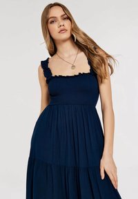 Marineblaues Kleid mit Rüschenträgern, gerafftem Oberteil und gestuftem Rock. Hergestellt aus glattem Stoff mit weicher Textur und einem fließenden Design.