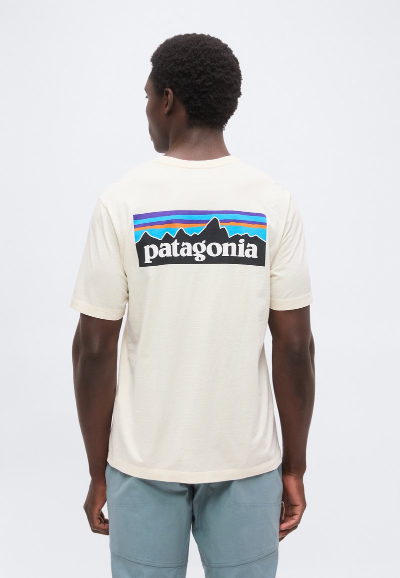 Άνδρας φοράει εκρού T-shirt Patagonia με πολύχρωμο λογότυπο βουνού στην πλάτη, συνδυασμένο με ανοιχτό μπλε παντελόνι, στέκεται μπροστά σε λευκό φόντο.