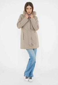 Beżowy płaszcz z futrzanym kołnierzem, o prostym kroju, z bocznymi kieszeniami i gładką fakturą, zestawiony z jasnoniebieskimi dżinsami i trampkami.